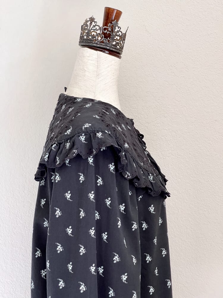 A black floral print historically inspired baroque blouse with large lace trimmed collar and long bishop sleeves is pictured on a mannequin from the side.