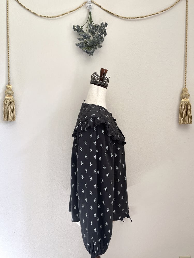 A black floral print historically inspired baroque blouse with large lace trimmed collar and long bishop sleeves is pictured on a mannequin from the side.