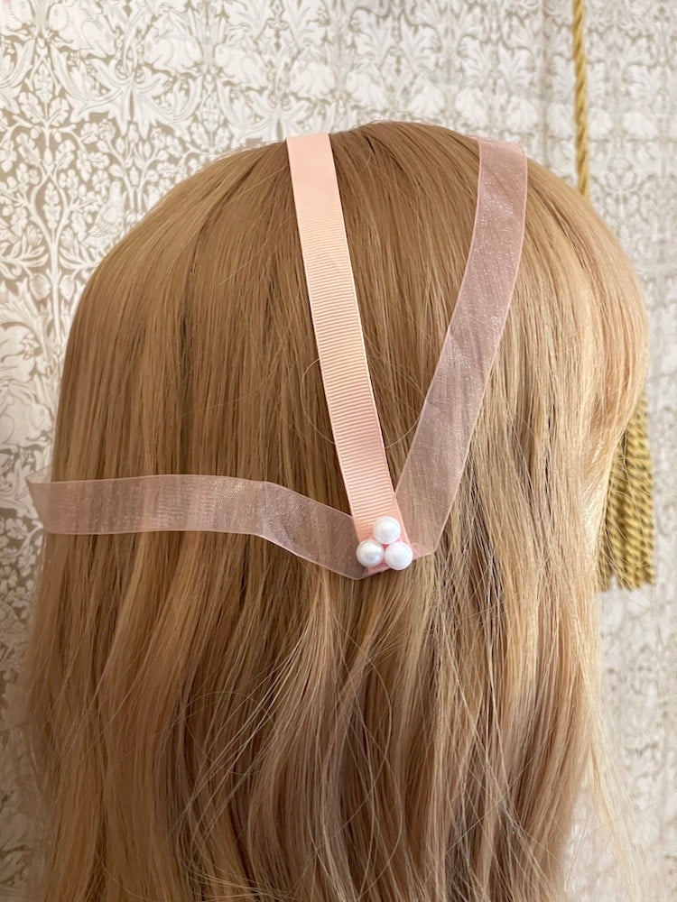 A handmade historically inspired regency era headdress in peachy pink with pearl accent is pictured on a mannequin.