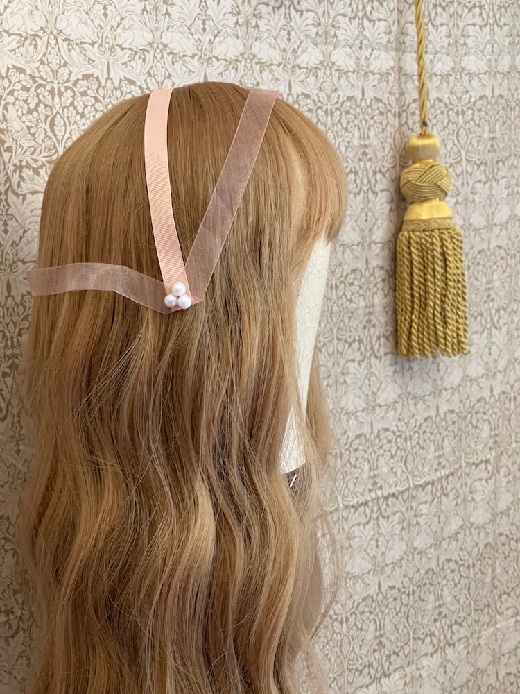 A handmade historically inspired regency era headdress in peachy pink with pearl accent is pictured on a mannequin.