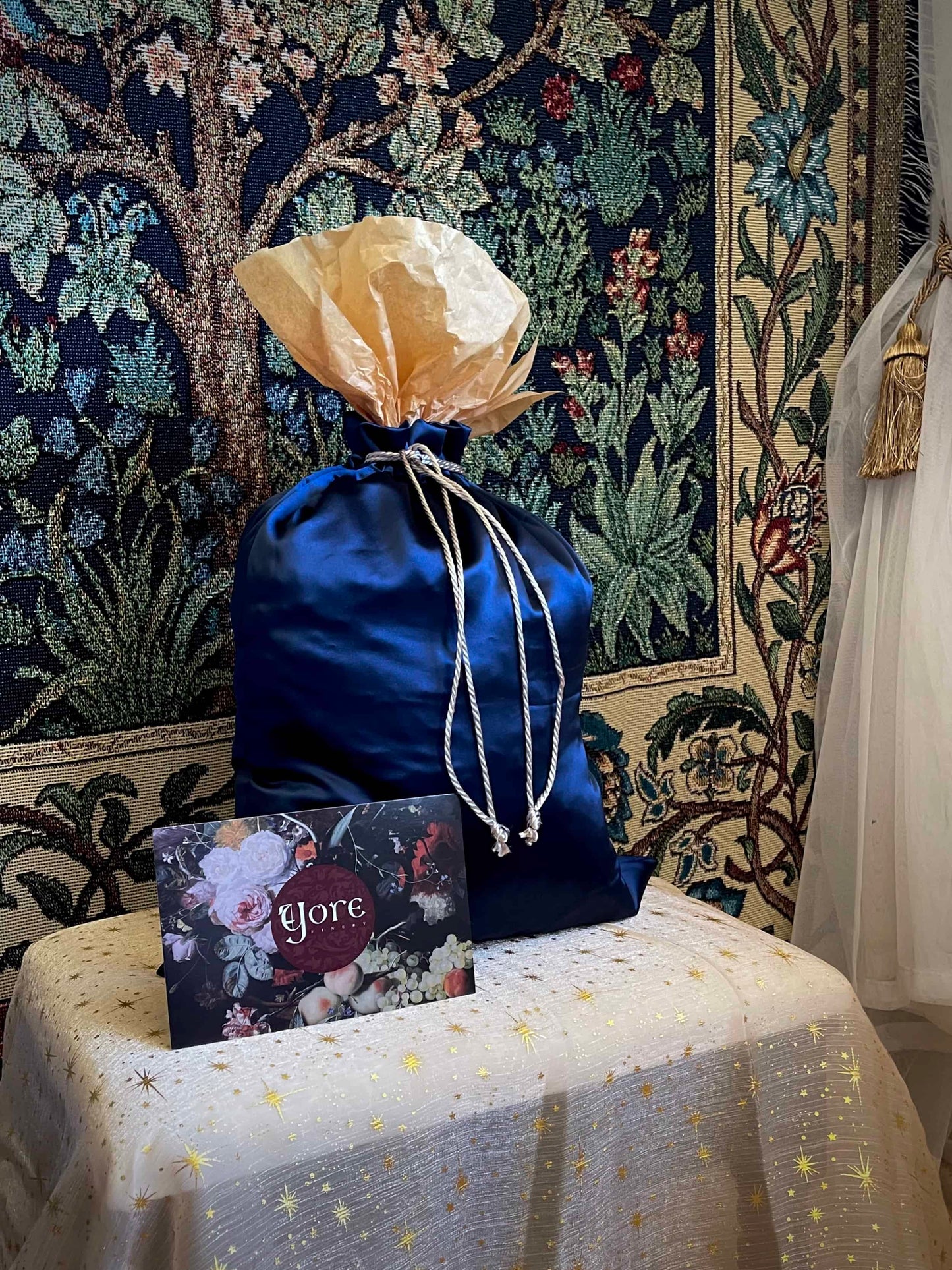 A historical fashion style bundle mystery bag in a satin drawstring bag sits on a celestial puff in front of a historically inspired tapestry.