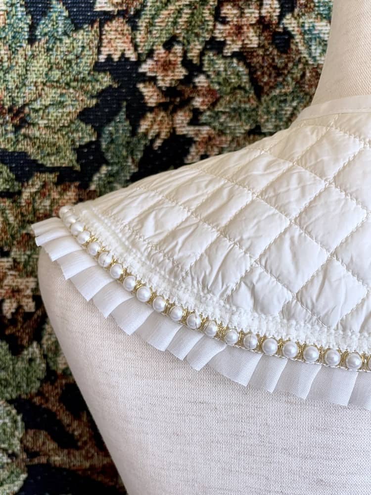 A Historically Inspired Quilted Round Collar with Ruffle & Pearl Trim in Ivory/White pictured on a mannequin in front of a historical tapestry.