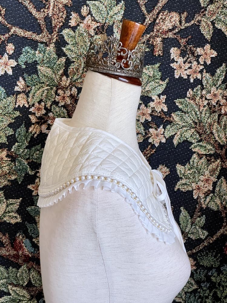 A Historically Inspired Quilted Round Collar with Ruffle & Pearl Trim in Ivory/White pictured on a mannequin in front of a historical tapestry.