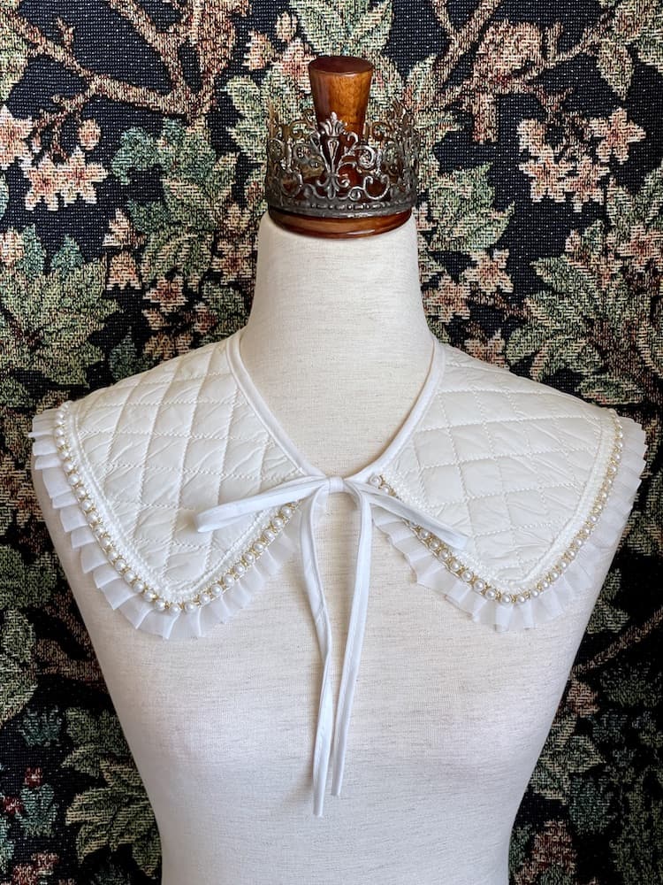 A Historically Inspired Quilted Round Collar with Ruffle & Pearl Trim in Ivory/White pictured on a mannequin in front of a historical tapestry.