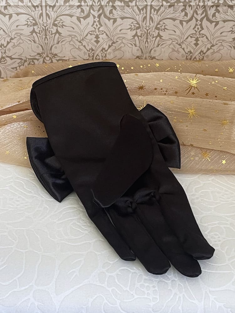 A pair of historically inspired Regency & Victorian era Satin Gloves with Large Bow Detail in Black, pictured on a historical backdrop.
