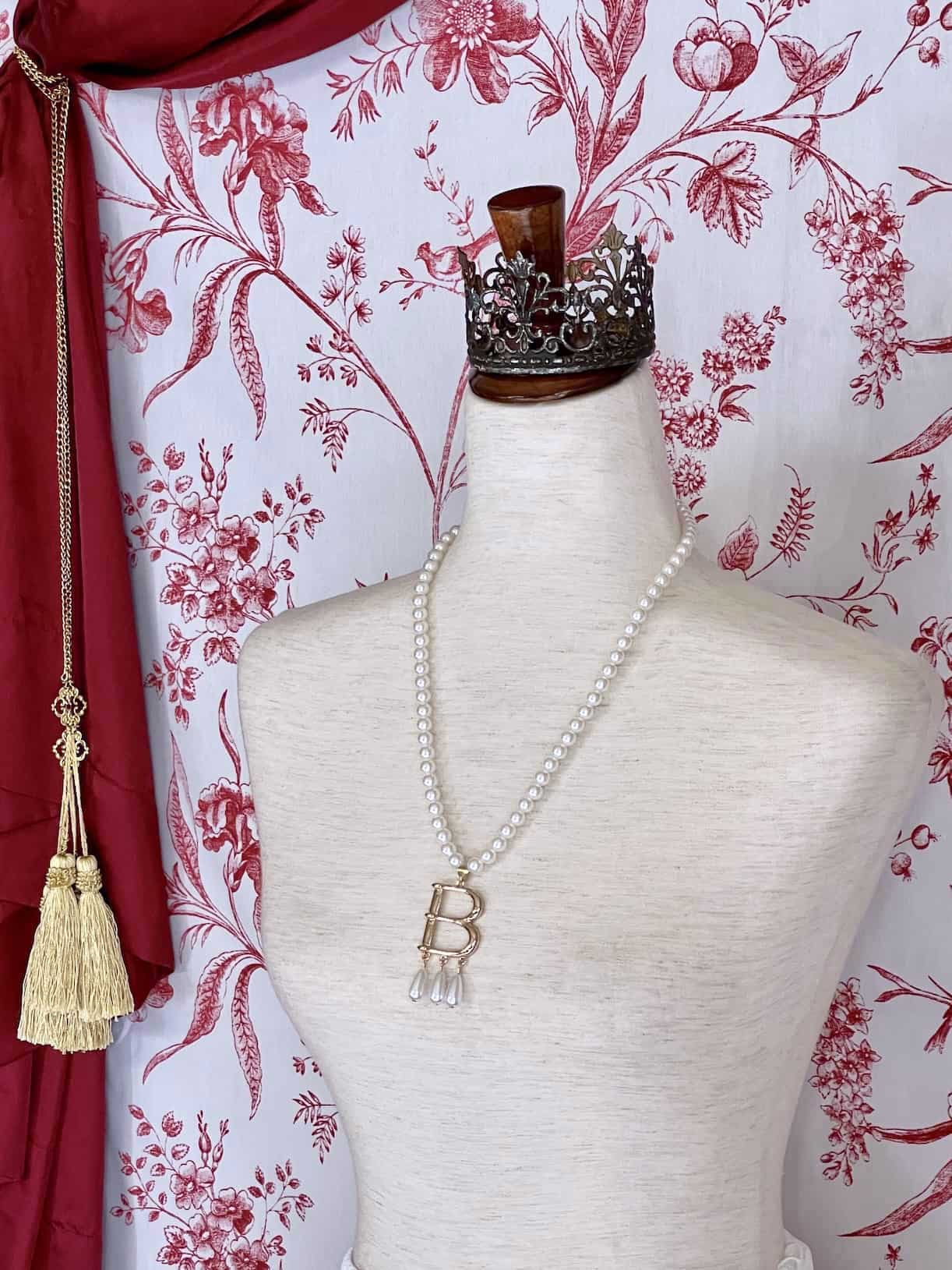 A Historical Tudor Fashion Inspired Anne Boleyn "B" Pearl Necklace on a mannequin in front of an ornate backdrop.