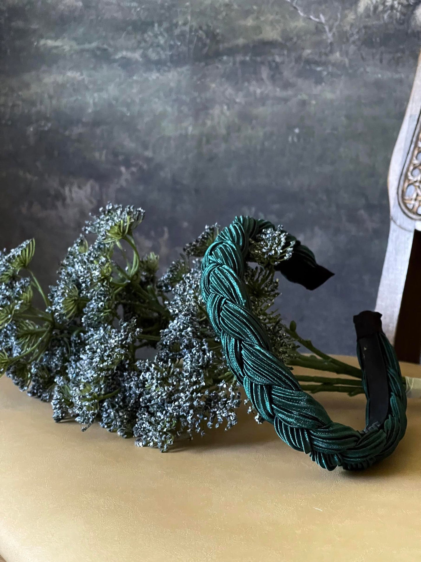 A historically inspired vintage style braided headband in dark forest green is pictured with a floral bouquet in front of an art history backdrop.