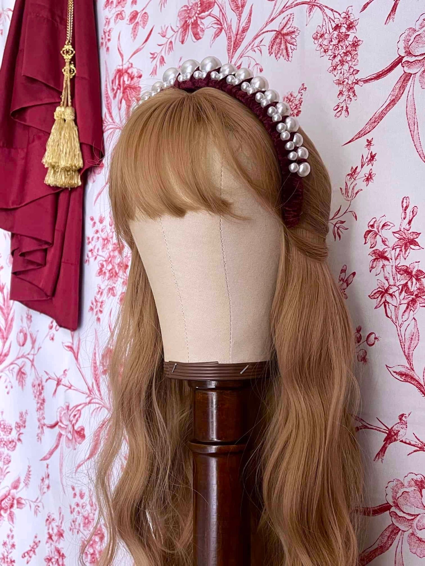 A Historically Inspired French Hood Style Velvet Pearl Headband in Burgundy in front of a toile backdrop.