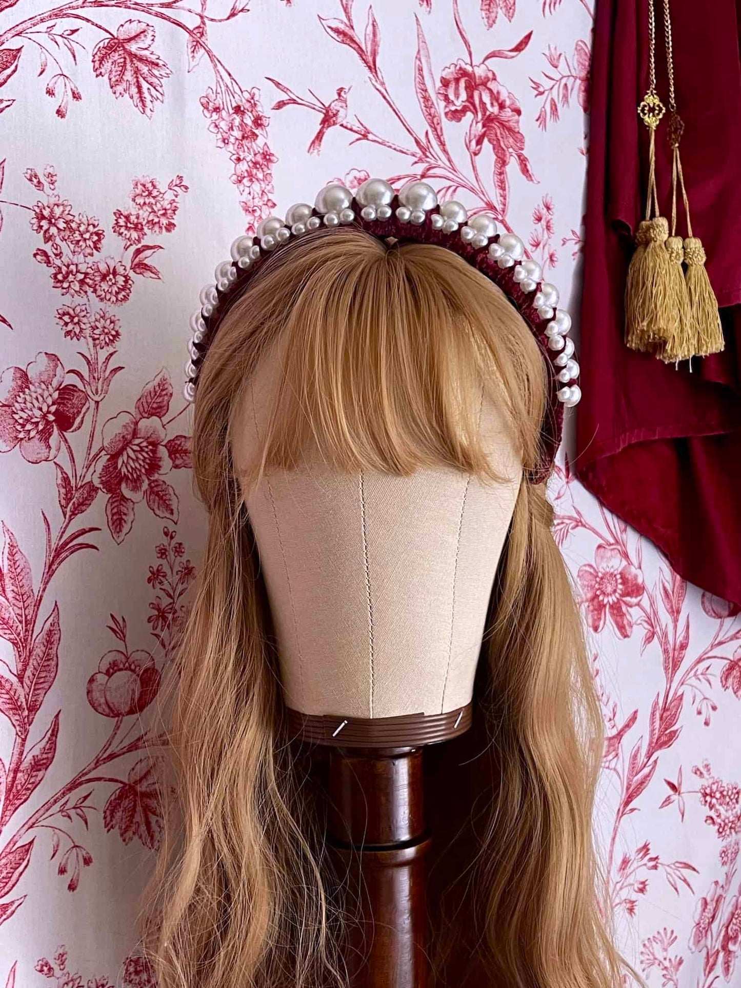 A Historically Inspired French Hood Style Velvet Pearl Headband in Burgundy in front of a toile backdrop.