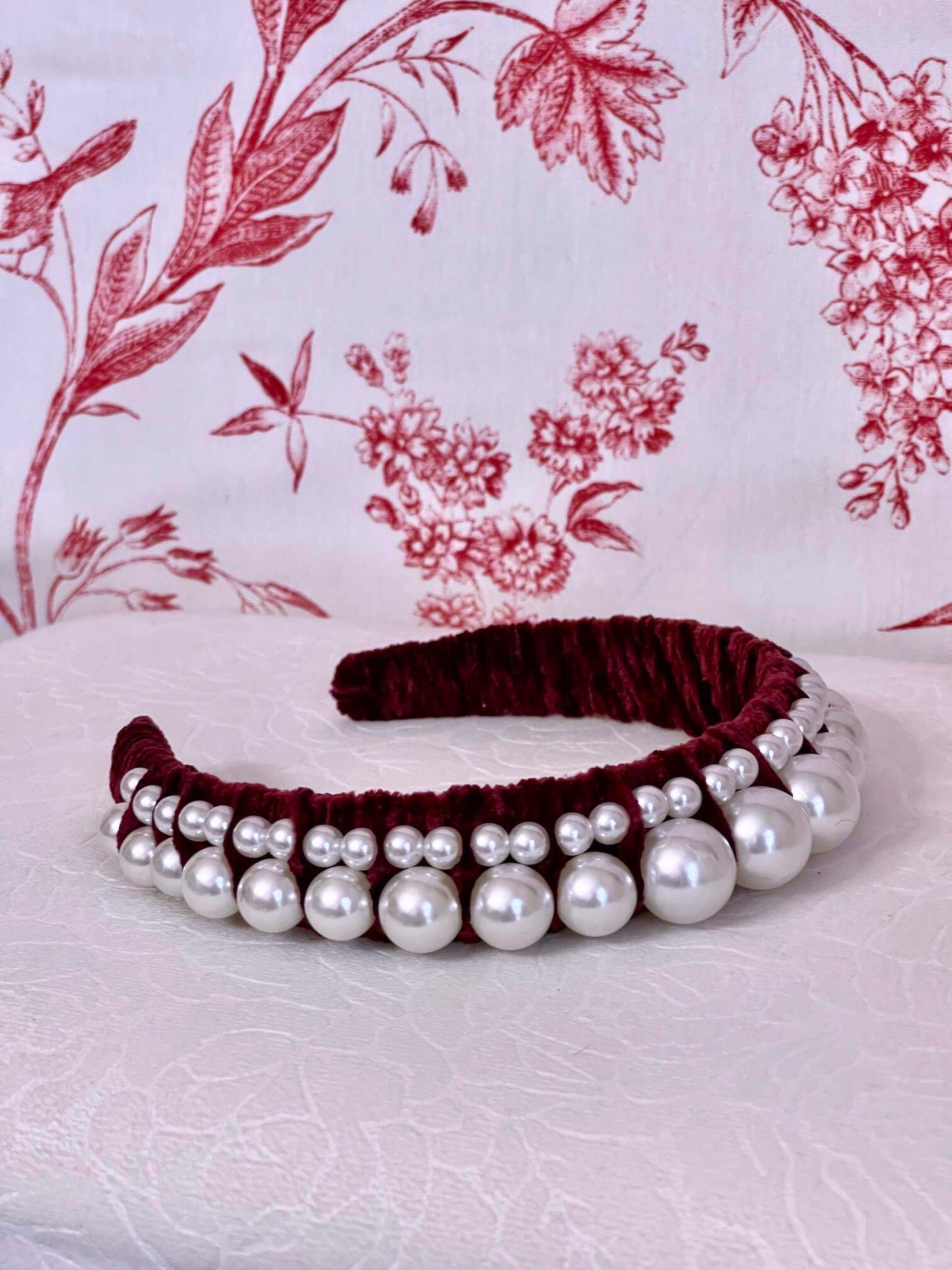 A Historically Inspired French Hood Style Velvet Pearl Headband in Burgundy in front of a toile backdrop.