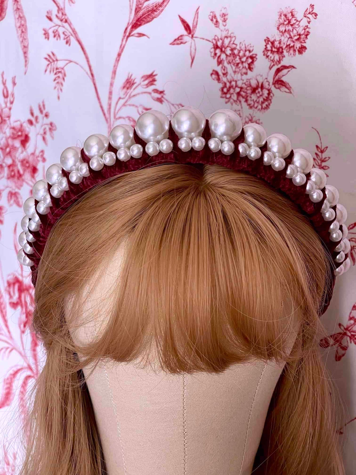 A Historically Inspired French Hood Style Velvet Pearl Headband in Burgundy in front of a toile backdrop.