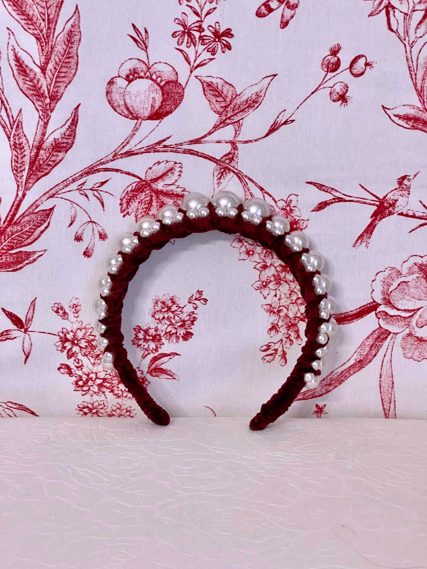 A Historically Inspired French Hood Style Velvet Pearl Headband in Burgundy in front of a toile backdrop.