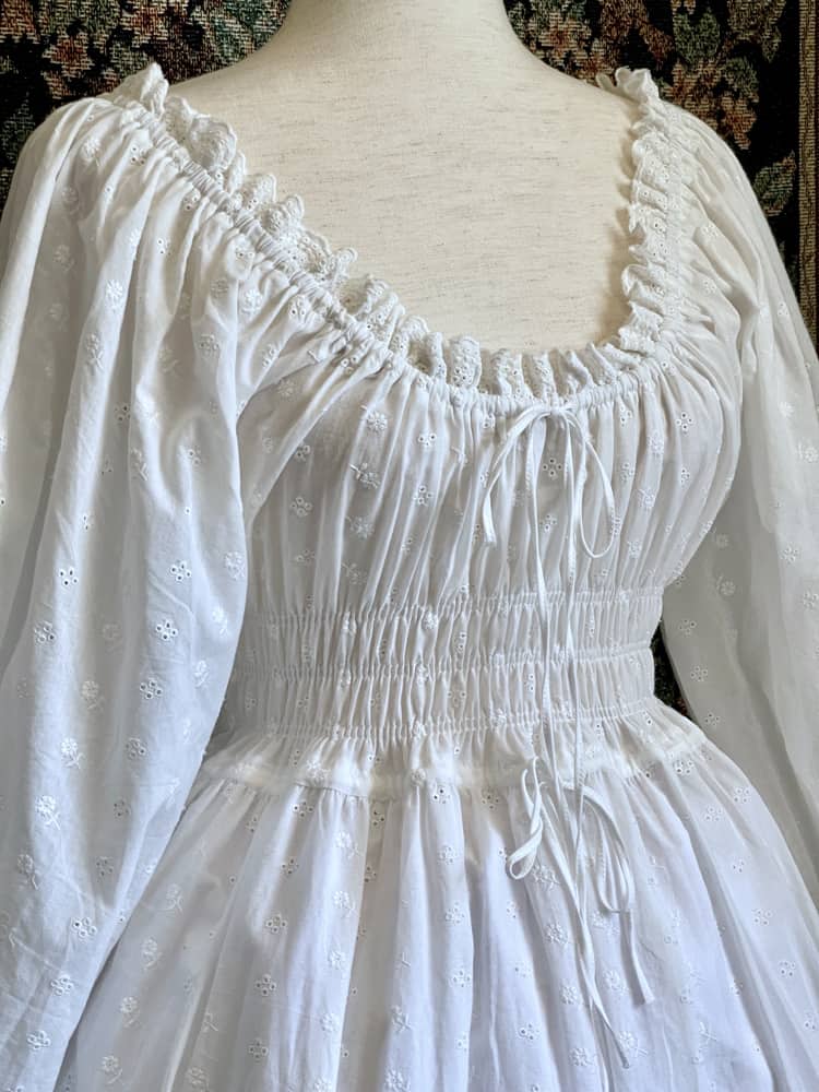 A Historically inspired white 100% cotton smocked chemise dress with bishop bell sleeves is pictured on a mannequin in front of an ornate tapestry background.