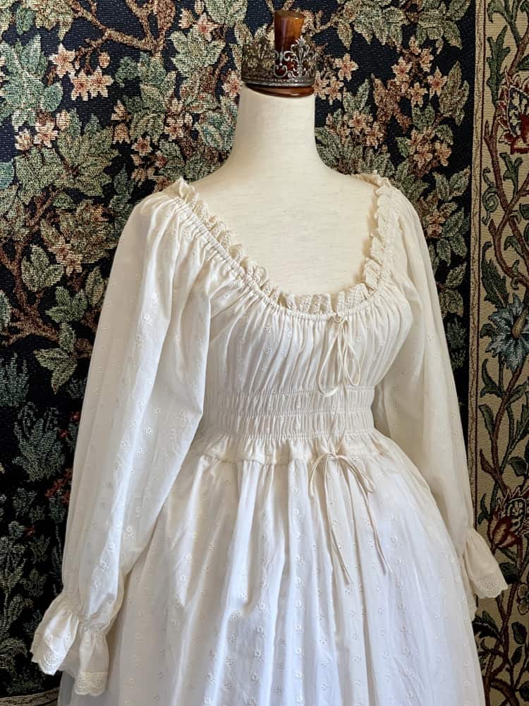 A Historically inspired sheer ivory 100% Cotton smocked chemise maxi dress with bishop bell sleeves is pictured on a mannequin in front of an ornate botanical tapestry.