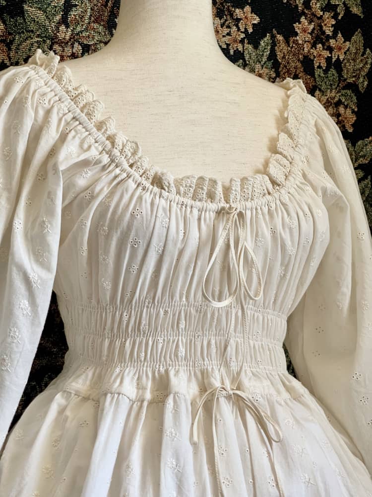 A Historically inspired sheer ivory 100% Cotton smocked chemise maxi dress with bishop bell sleeves is pictured on a mannequin in front of an ornate botanical tapestry.