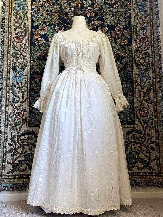 A Historically inspired sheer ivory 100% Cotton smocked chemise maxi dress with bishop bell sleeves is pictured on a mannequin in front of an ornate botanical tapestry.