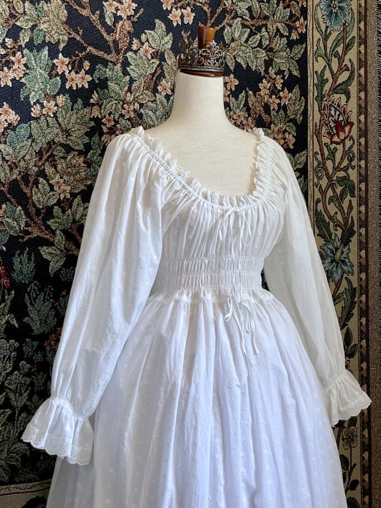 A Historically inspired white 100% cotton smocked chemise dress with bishop bell sleeves is pictured on a mannequin in front of an ornate tapestry background.