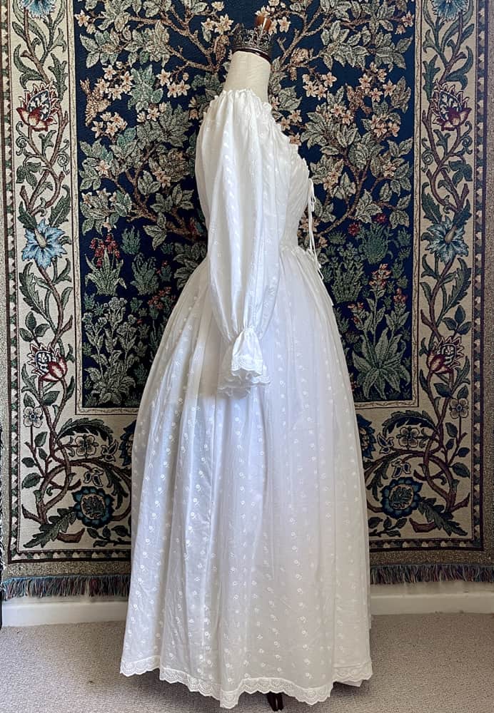 A Historically inspired white 100% cotton smocked chemise dress with bishop bell sleeves is pictured on a mannequin in front of an ornate tapestry background.
