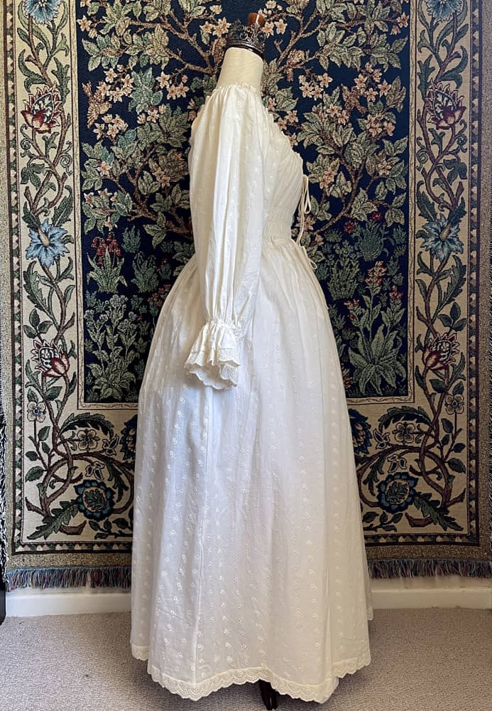 A Historically inspired sheer ivory 100% Cotton smocked chemise maxi dress with bishop bell sleeves is pictured on a mannequin in front of an ornate botanical tapestry.