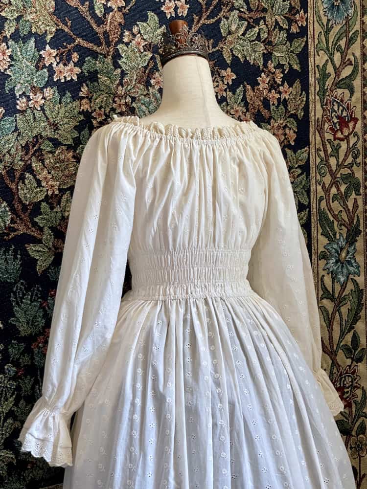 A Historically inspired sheer ivory 100% Cotton smocked chemise maxi dress with bishop bell sleeves is pictured on a mannequin in front of an ornate botanical tapestry.