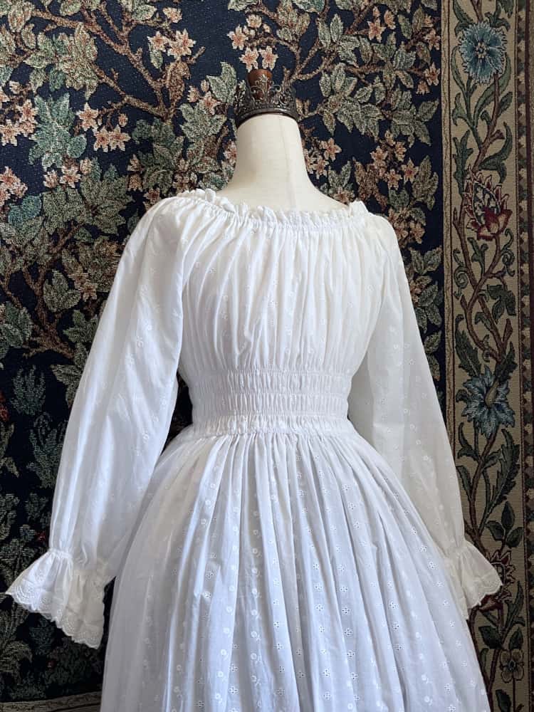 A Historically inspired white 100% cotton smocked chemise dress with bishop bell sleeves is pictured on a mannequin in front of an ornate tapestry background.