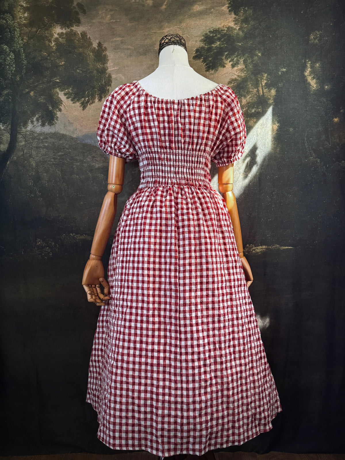 A red and white gingham midi dress with puff sleeves and a smocked waist, displayed on a mannequin, set against a dark vintage-style landscape backdrop.