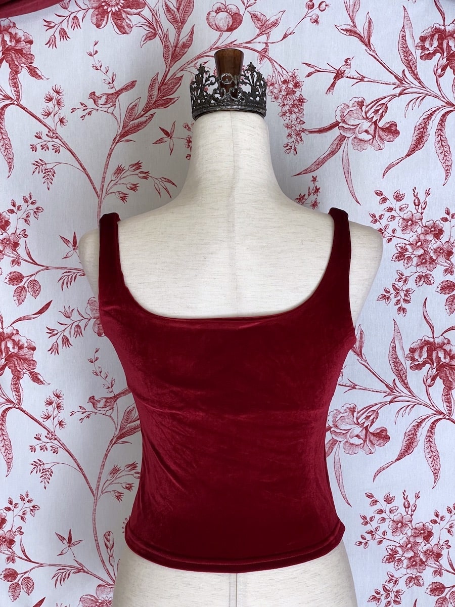 A historically inspired burgundy red velvet ruched milkmaid bustier top is pictured on a mannequin in front of a red and white ornate toile Wallpaper.