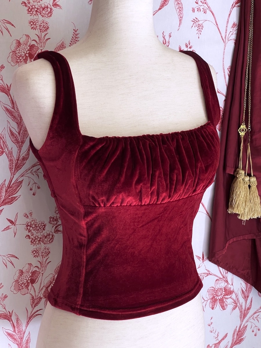A historically inspired burgundy red velvet ruched milkmaid bustier top is pictured on a mannequin in front of a red and white ornate toile Wallpaper.