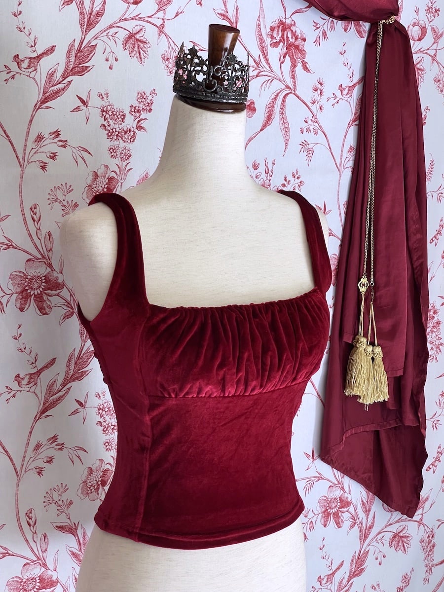 A historically inspired burgundy red velvet ruched milkmaid bustier top is pictured on a mannequin in front of a red and white ornate toile Wallpaper.