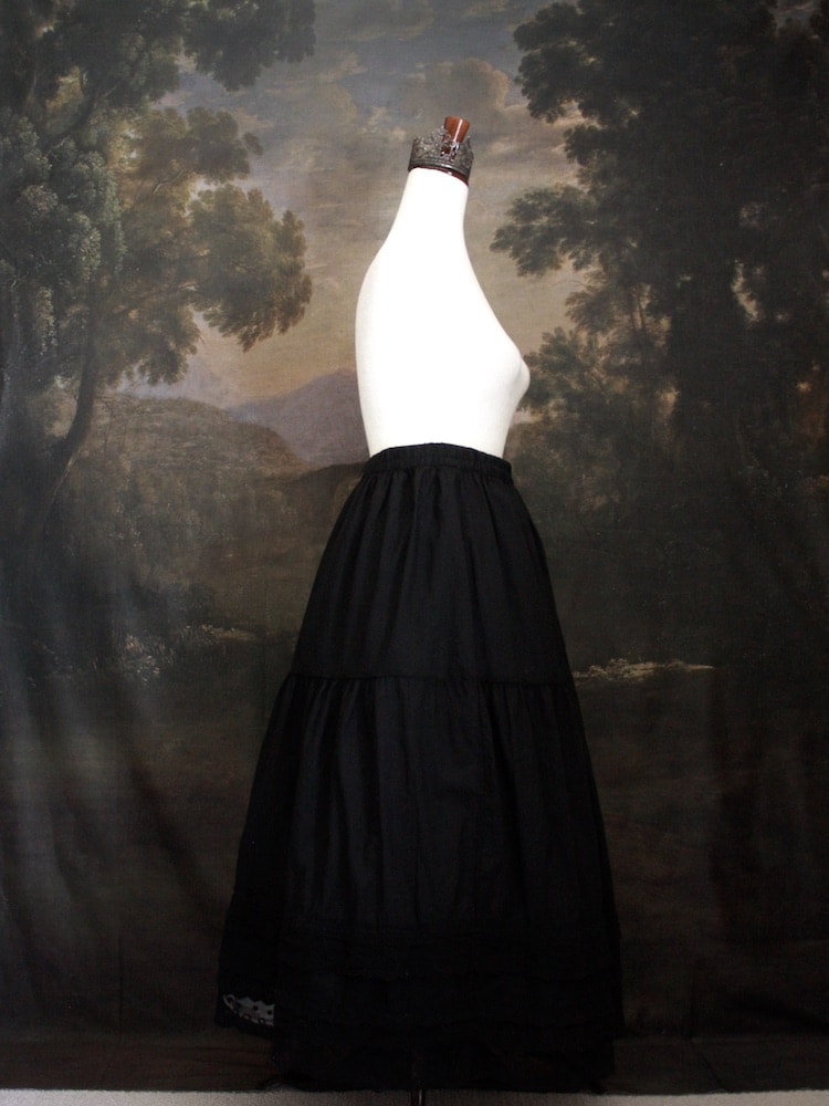 A Historically Inspired bohemian Tiered Cotton Maxi Skirt with floral Lace Trim in Black on a mannequin in front of an art history backdrop.