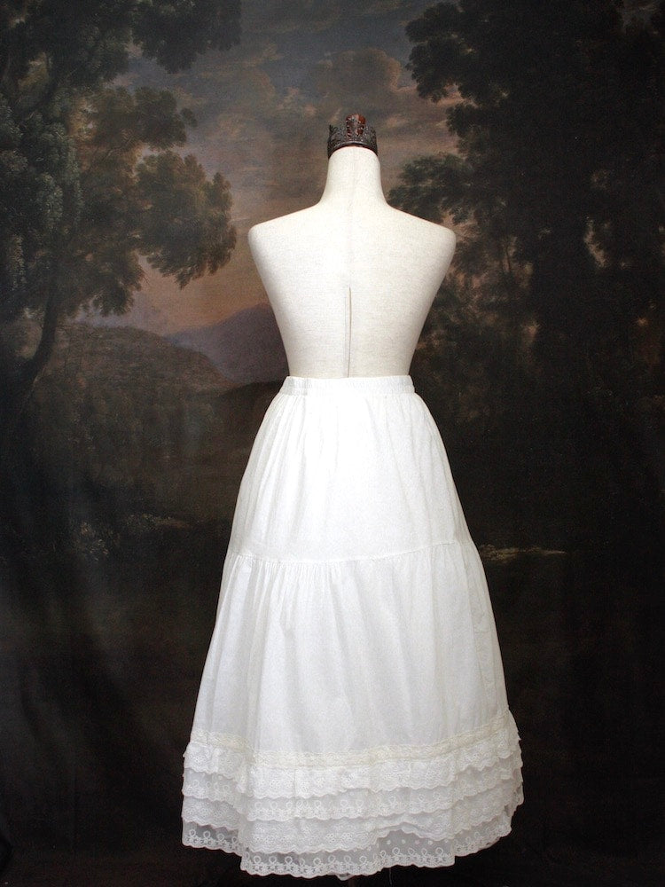 A Historically Inspired bohemian Tiered Cotton Maxi Skirt with floral Lace Trim in White on a mannequin in front of an art history backdrop.