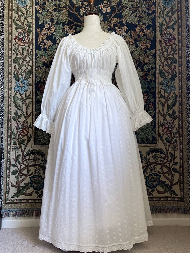A Historically inspired white 100% cotton smocked chemise dress with bishop bell sleeves is pictured on a mannequin in front of an ornate tapestry background.