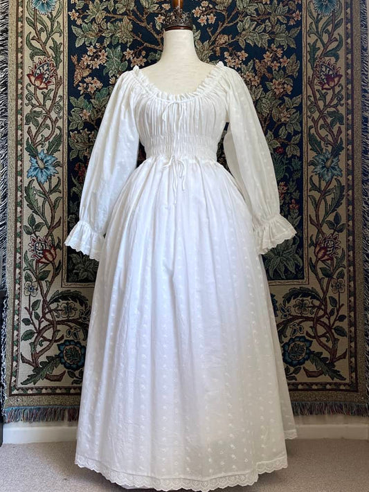 A Historically inspired white 100% cotton smocked chemise dress with bishop bell sleeves is pictured on a mannequin in front of an ornate tapestry background.