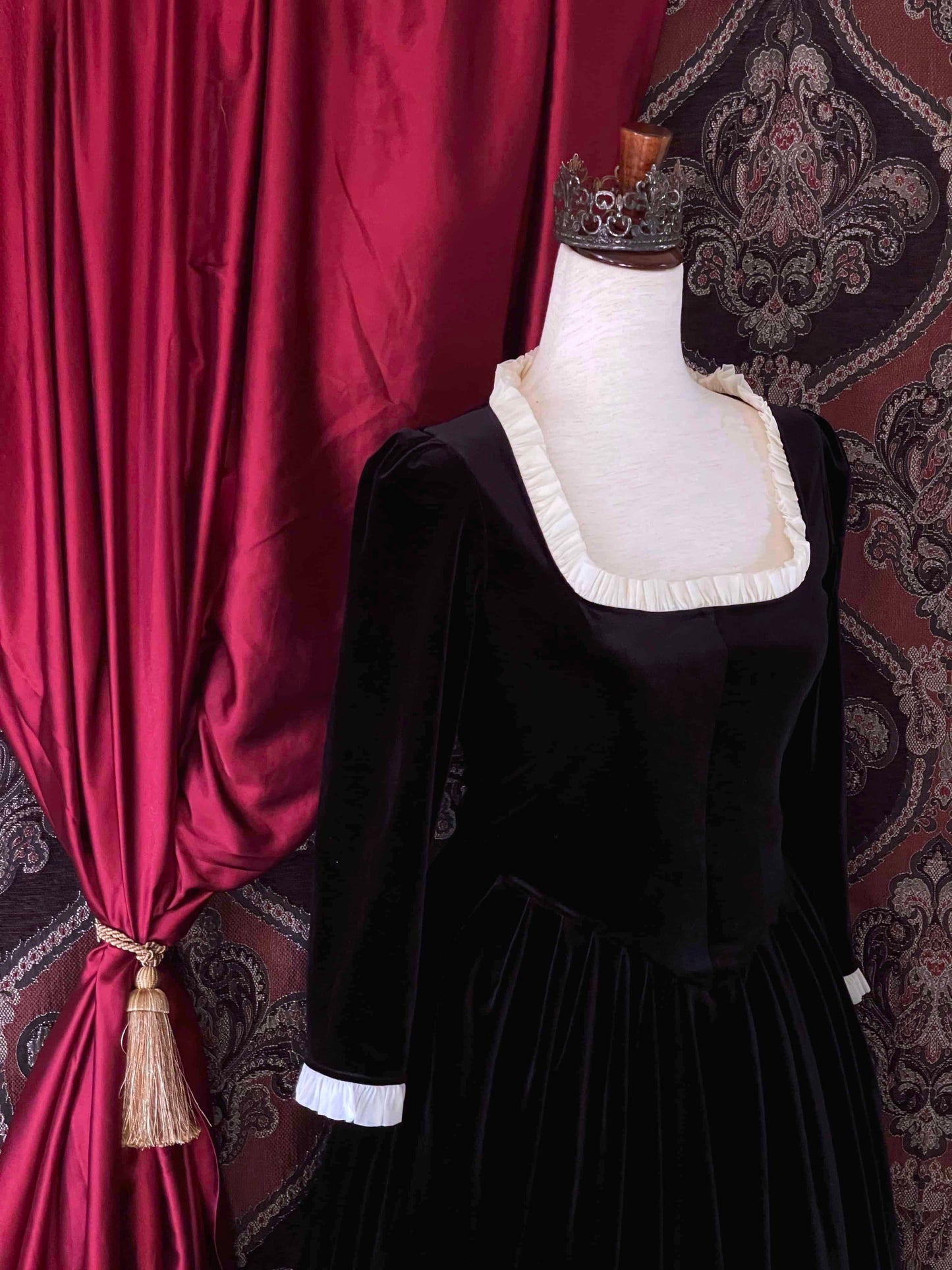 A historically inspired renaissance tudor era gothic black velvet gown with structured corset style bodice and ruffle lace trim, pictured on a mannequin in front of an ornate background.