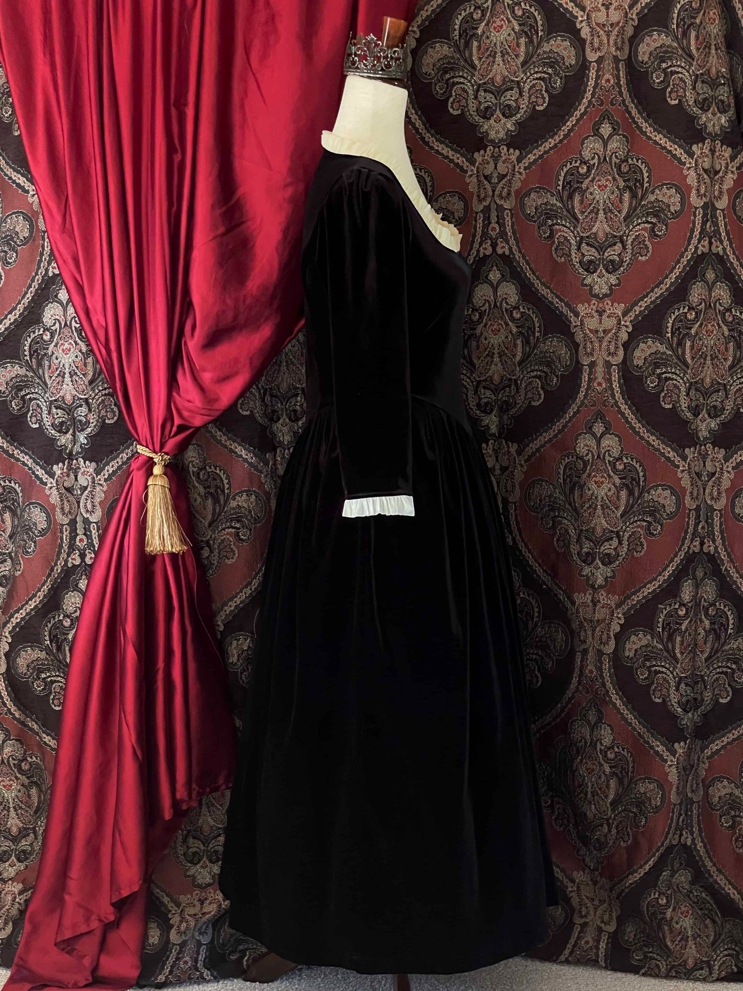 A historically inspired renaissance tudor era gothic black velvet gown with structured corset style bodice and ruffle lace trim, pictured on a mannequin in front of an ornate background.