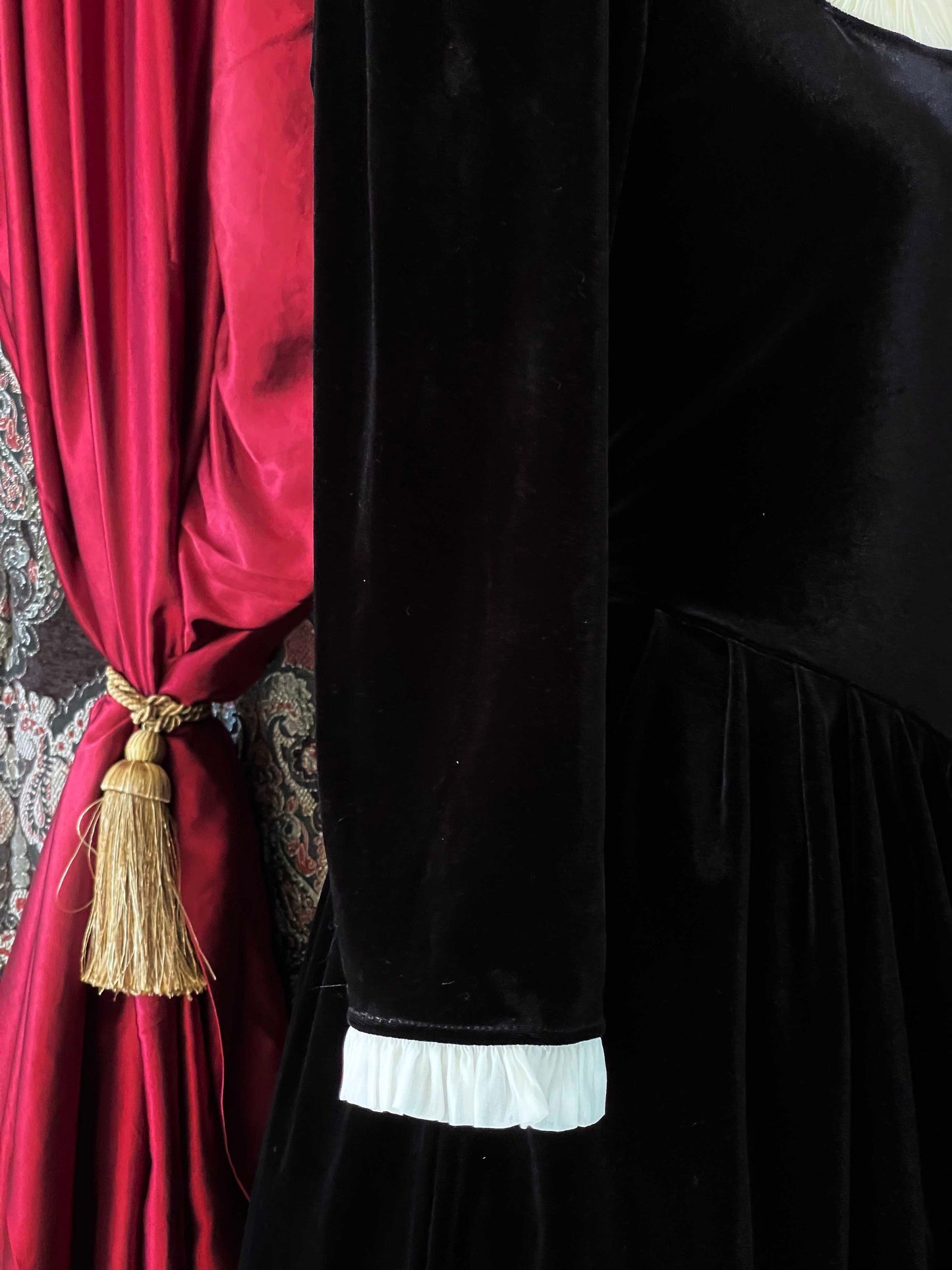 A historically inspired renaissance tudor era gothic black velvet gown with structured corset style bodice and ruffle lace trim, pictured on a mannequin in front of an ornate background.