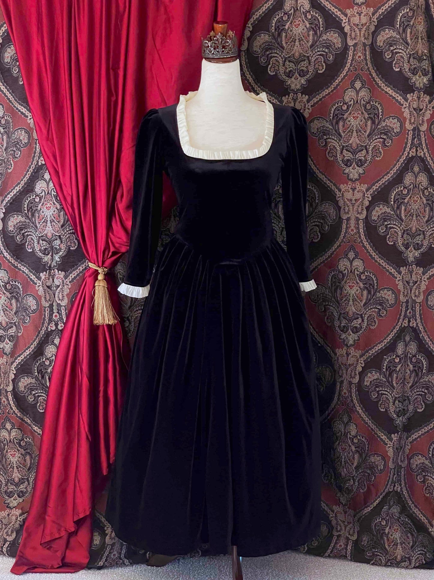 A historically inspired renaissance tudor era gothic black velvet gown with structured corset style bodice and ruffle lace trim, pictured on a mannequin in front of an ornate background.