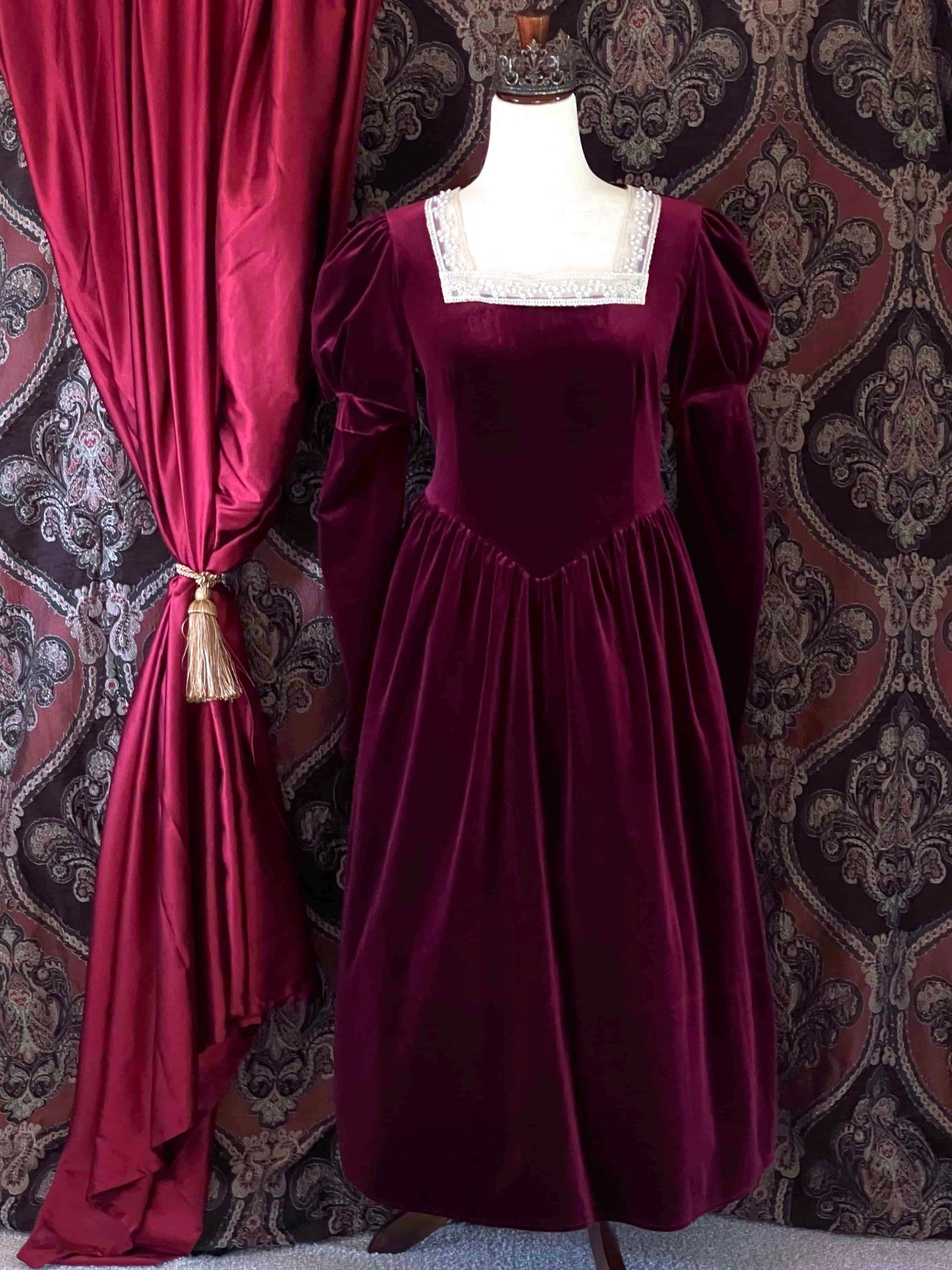 A Renaissance and Tudor inspired Burgundy Velvet Juliet Sleeve Gown with lace trim is pictured on a mannequin in front of a historical ornate backdrop.