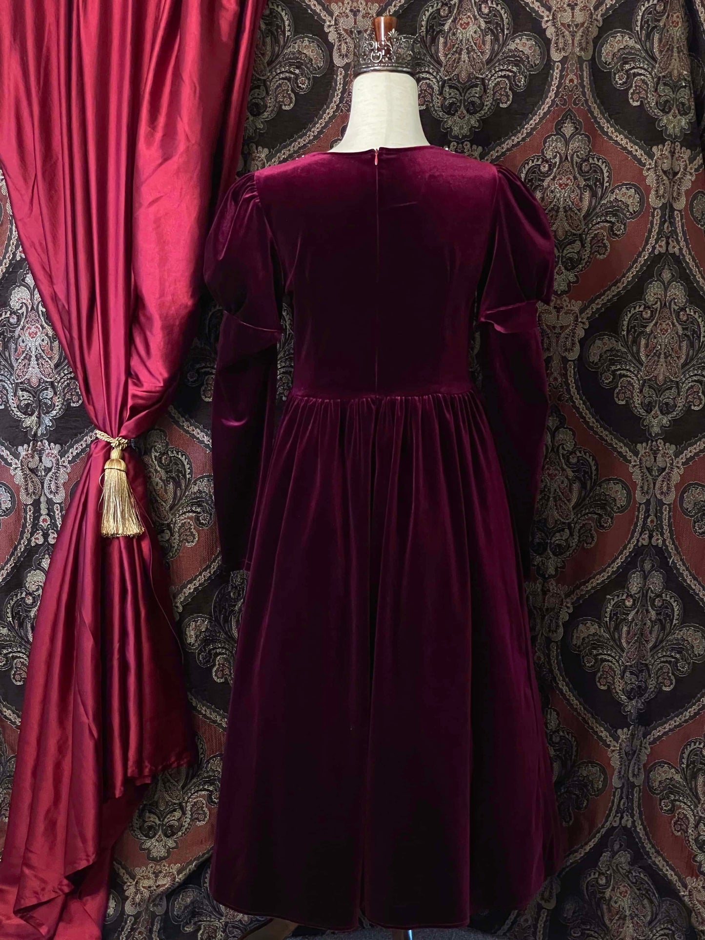 A Renaissance and Tudor inspired Burgundy Velvet Juliet Sleeve Gown with lace trim is pictured on a mannequin in front of a historical ornate backdrop.