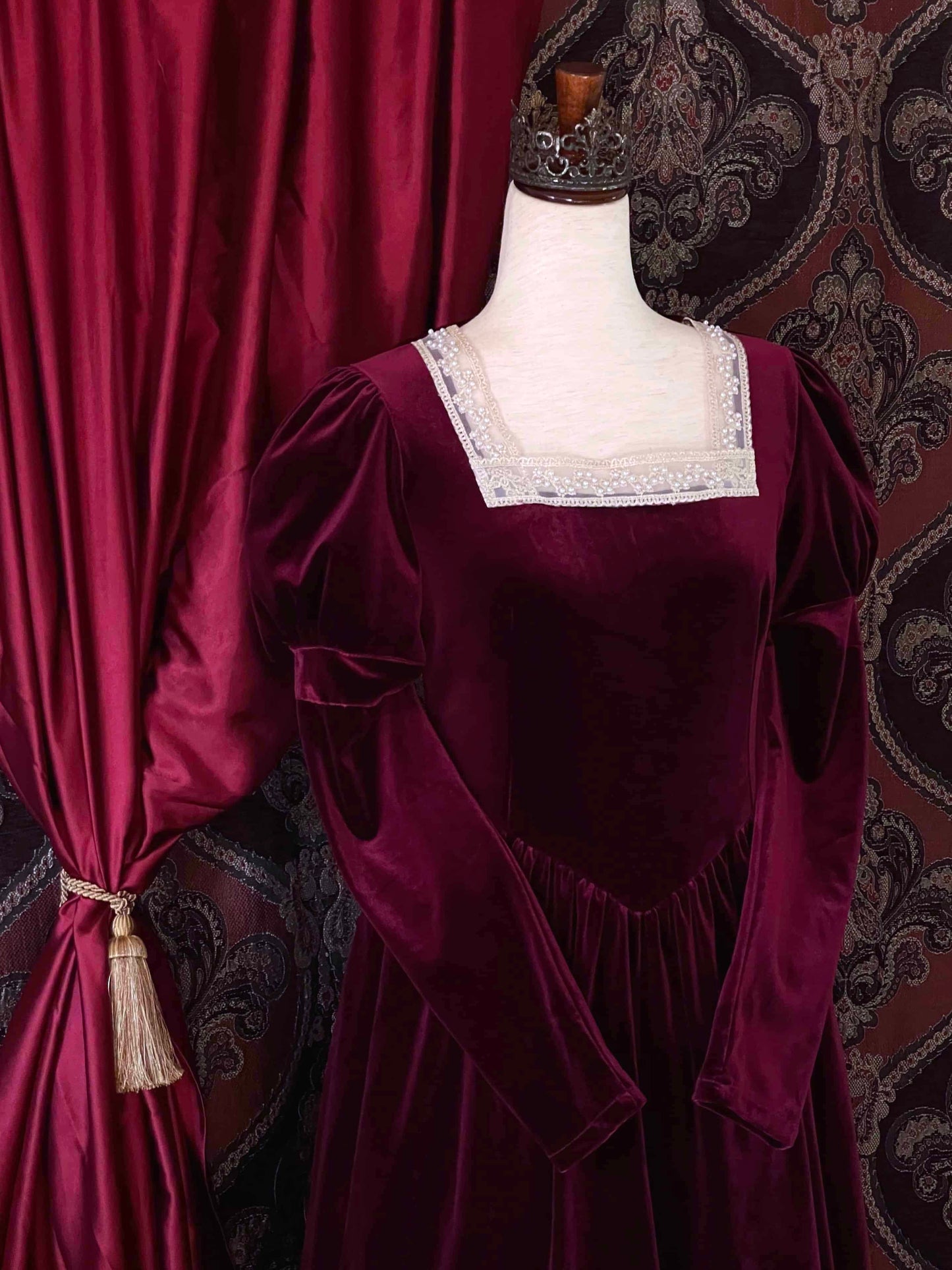 A Renaissance and Tudor inspired Burgundy Velvet Juliet Sleeve Gown with lace trim is pictured on a mannequin in front of a historical ornate backdrop.