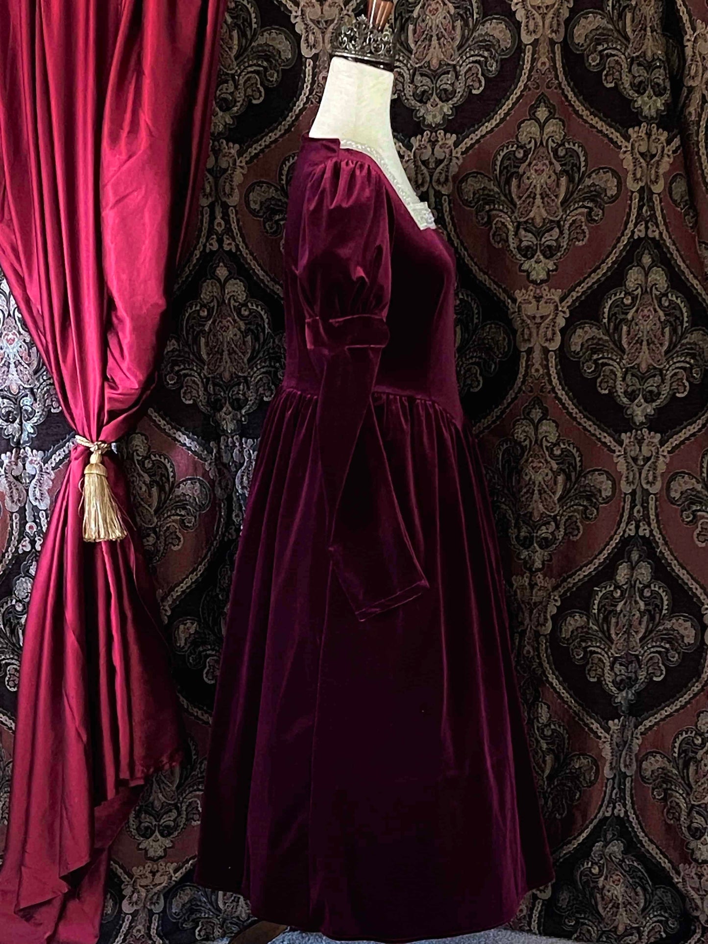 A Renaissance and Tudor inspired Burgundy Velvet Juliet Sleeve Gown with lace trim is pictured on a mannequin in front of a historical ornate backdrop.