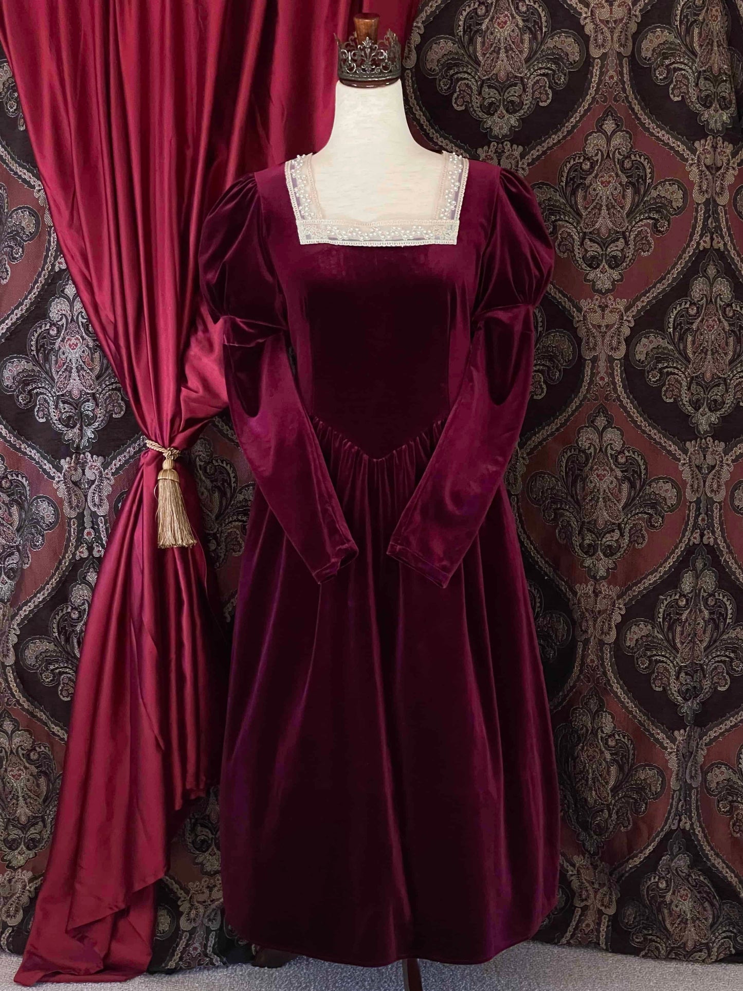 A Renaissance and Tudor inspired Burgundy Velvet Juliet Sleeve Gown with lace trim is pictured on a mannequin in front of a historical ornate backdrop.