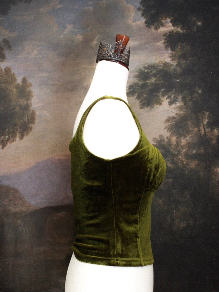 A Historically Inspired medieval fantasy Velvet Milkmaid Bustier Top in Moss Green in front of a historical painting backdrop of a forest.