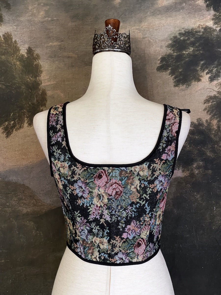 A historically inspired black floral print woven gobelin tapestry corset is pictured on a mannequin in front of an ornate landscape painting wallpaper.