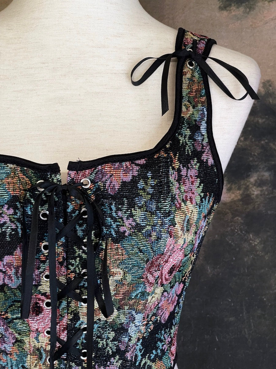 A historically inspired black floral print woven gobelin tapestry corset is pictured on a mannequin in front of an ornate landscape painting wallpaper.