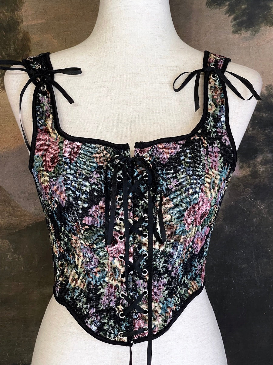 A historically inspired black floral print woven gobelin tapestry corset is pictured on a mannequin in front of an ornate landscape painting wallpaper.