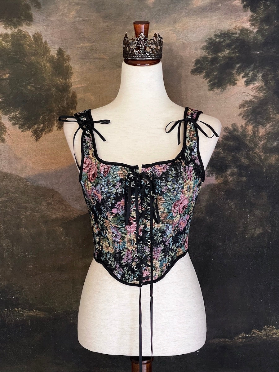 A historically inspired black floral print woven gobelin tapestry corset is pictured on a mannequin in front of an ornate landscape painting wallpaper.