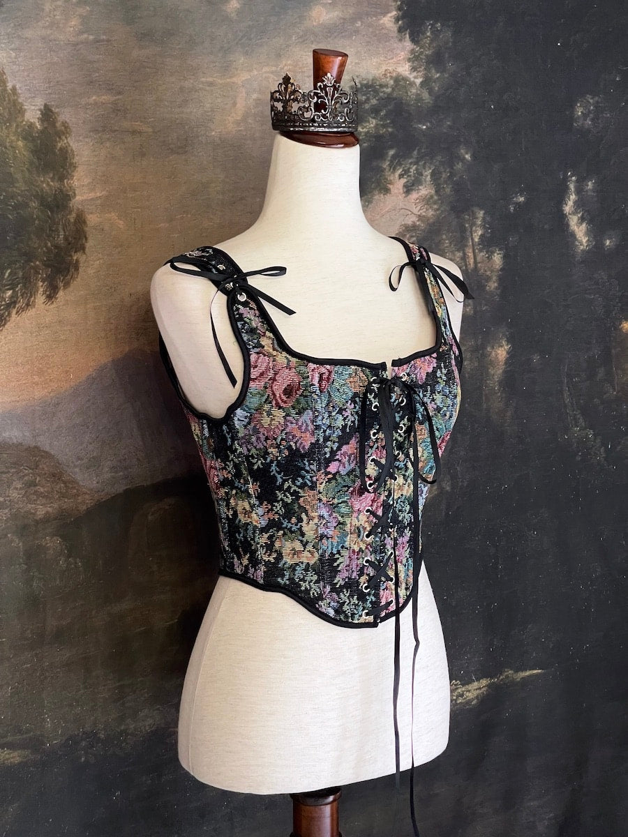 A historically inspired black floral print woven gobelin tapestry corset is pictured on a mannequin in front of an ornate landscape painting wallpaper.