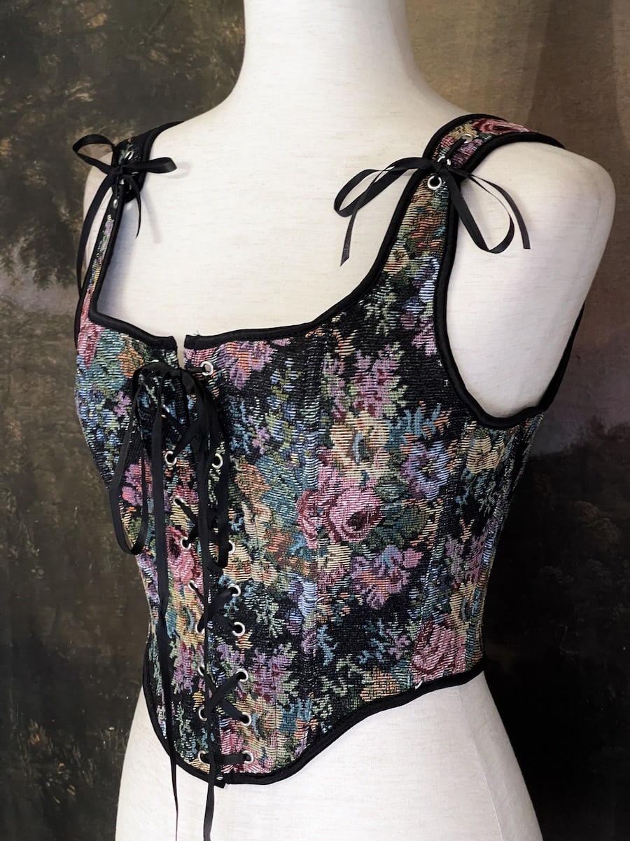 A historically inspired black floral print woven gobelin tapestry corset is pictured on a mannequin in front of an ornate landscape painting wallpaper.