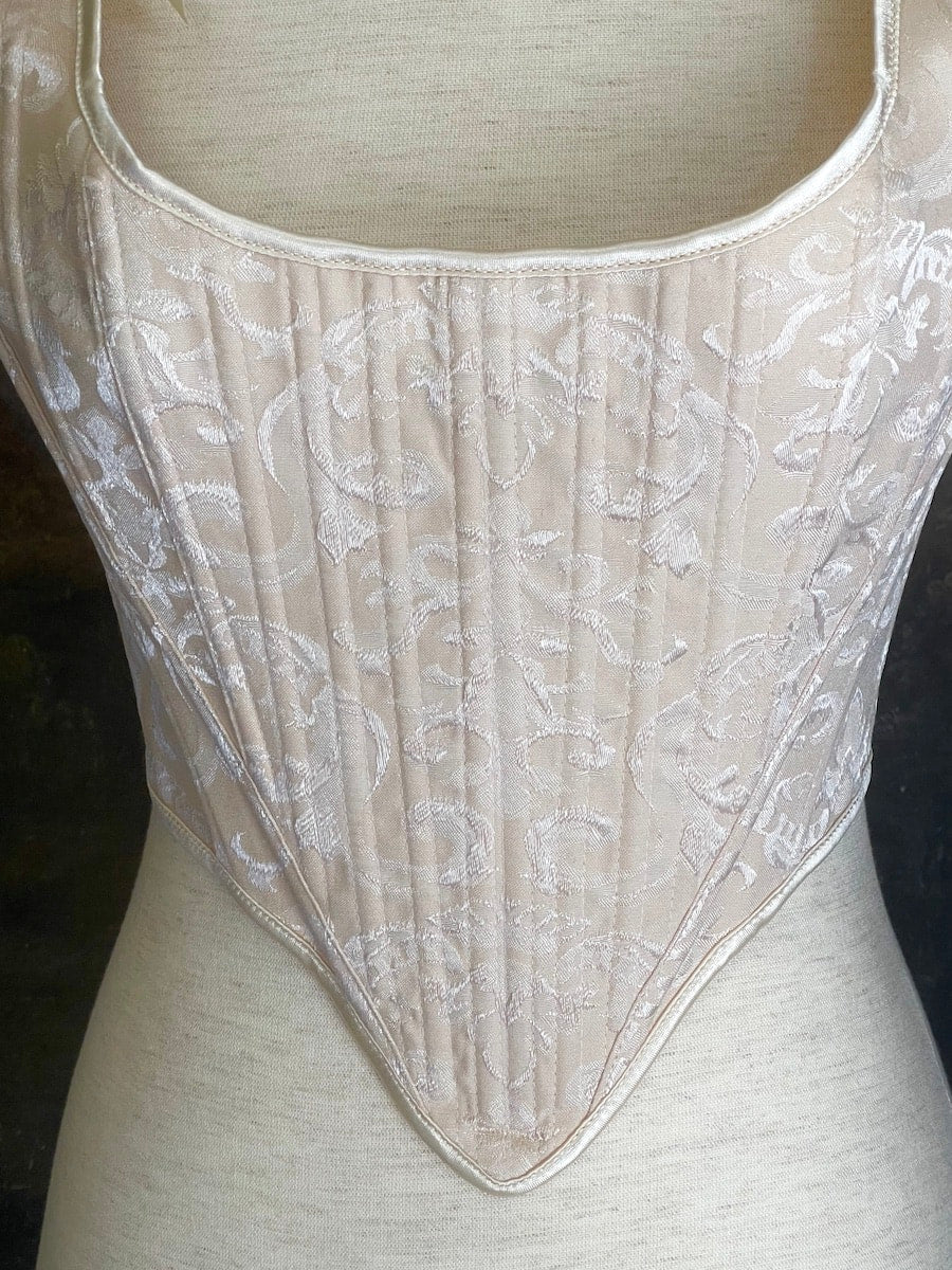 A Historically inspired Tudor, baroque, or renaissance era corset with champagne-ivory colored ornate jacquard fabric is pictured on a mannequin in front of a historic painted landscape backdrop.